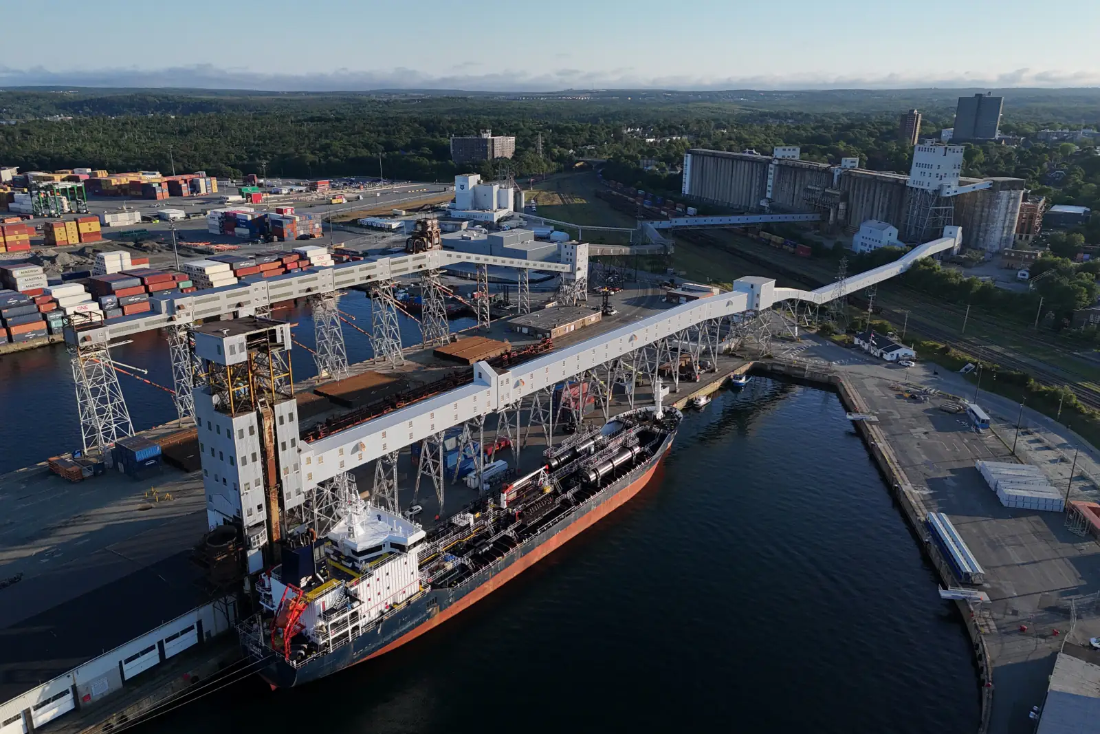 Aerial view of a bulk carrier berthed beneath the loading galleries of a grain terminal, with silos and a container yard behind it.