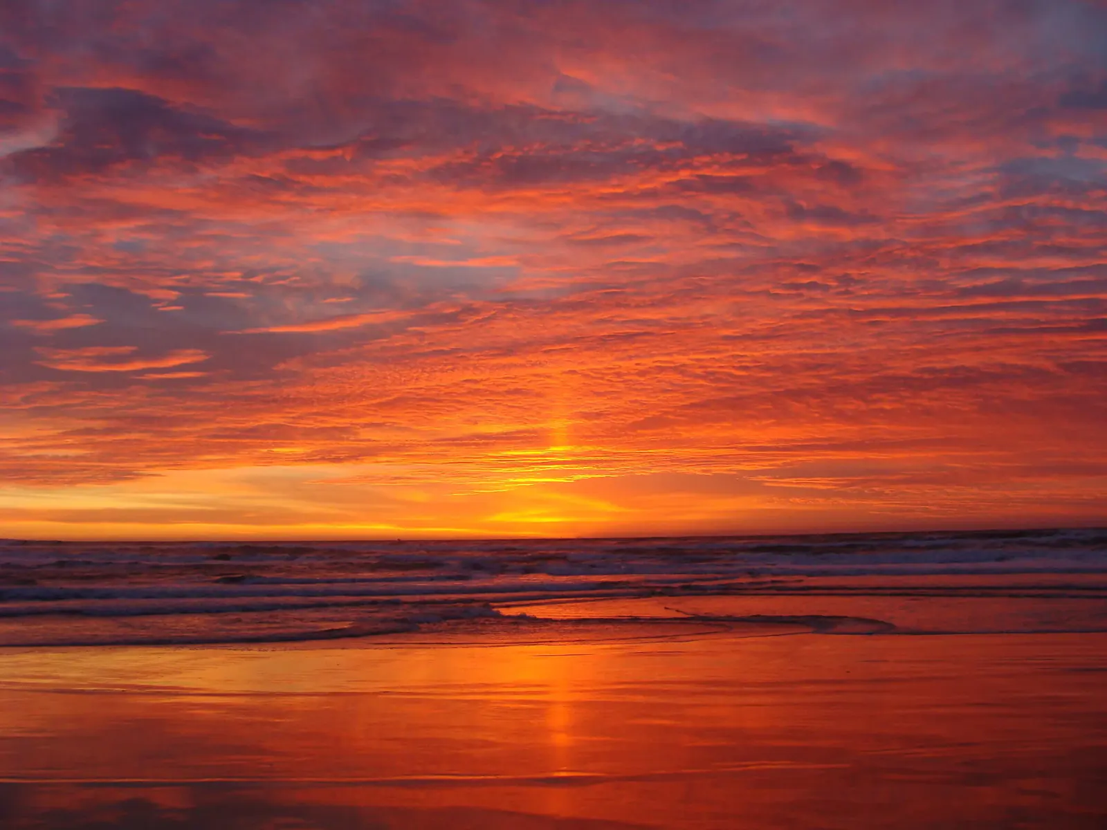 Sunset over the Pacific Ocean, with surf breaking on a wide beach under a red and gold sky.